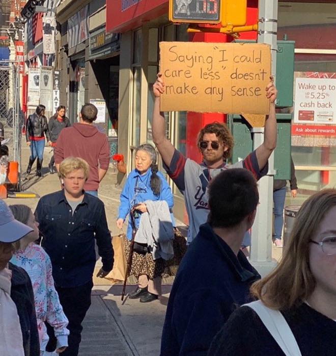 The meme is a photo of a man holding a sign on a street corner. The sign reads: "Saying 'I can care less' doesn't make any sense." The man is likely a protestor or a street performer, and the sign is a humorous commentary on the common phrase "I could care less," which is often used to express a lack of concern or interest.